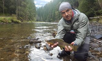 A fisherman holds a large trout in his hands. In the background, a clear river flows, surrounded by green trees.