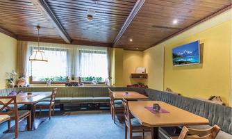 An inviting restaurant with wood-paneled ceiling and cozy seating areas. The walls are painted in a warm yellow tone and a television screen displays a mountain landscape.