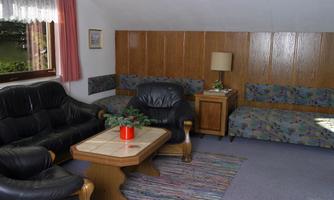 A cozy living room with a black leather sofa set and a wooden table. The walls are paneled with wood and there are colorful upholstered chairs.