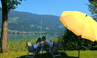 A couple sits under a yellow sun umbrella and enjoys the view of a lake and the mountains. The surroundings are green and inviting.