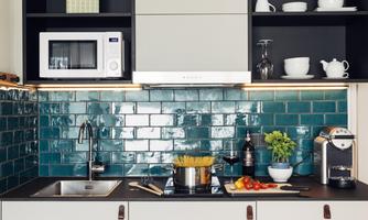 A modern kitchen with blue tiles and a microwave. On the countertop are a pot of pasta, fresh vegetables, and a coffee machine.