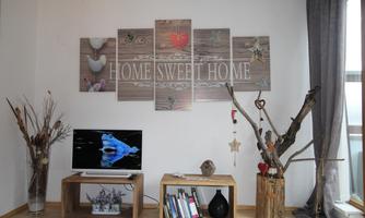 A cozy living room with a modern TV corner and decorative wooden furniture. On the wall hangs a piece of art with the inscription "HOME SWEET HOME".