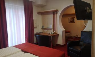 A comfortable hotel room with two beds, a desk, and a television. The room has large windows with curtains in vibrant red.