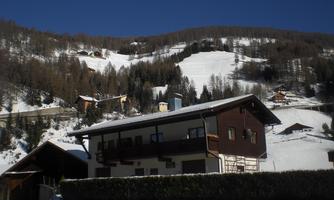 A cozy wooden house in a snowy landscape. In the background, forested hills and ski areas can be seen.