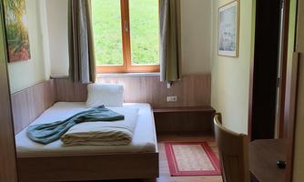 A simple and cozy room with a bed, a table, and a window overlooking a green landscape. The walls are light, and the furnishings are functional.