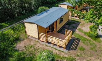 A modern wooden cabin with a porch in a green environment. The cabin is surrounded by trees and meadows.