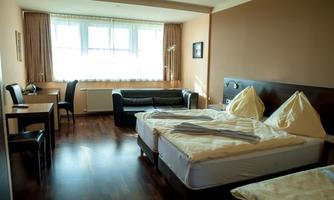 A simple room with two beds, a desk, and a sofa. The room has large windows and a warm color tone.