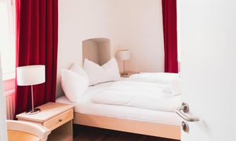 A bright bedroom with a double bed and red curtains. Next to the bed are bedside lamps and a small table.