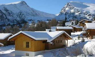 A snowy landscape with wooden chalets and majestic mountains in the background. The sky is clear and blue.