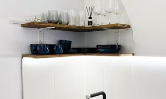 A modern kitchen with a black sink and an elegant faucet. Above, glasses and dishes are on an open wooden shelf.