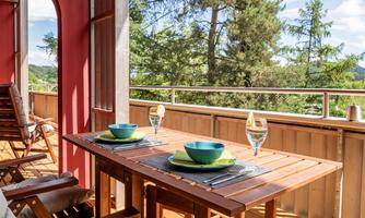 A cozy balcony with wooden furniture and a table set for two. Surrounded by views of trees and a clear sky.
