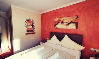 A cozy bedroom with a large bed and decorative pillows. The orange wall is decorated with a piece of art depicting hands.