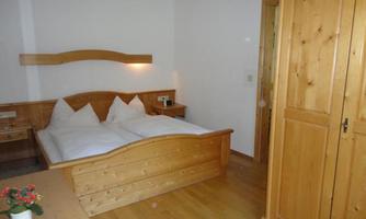 A simple bedroom with a wooden double bed and two comfortable pillows. There is a wooden wardrobe and a small table with a flower.