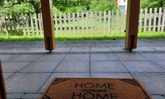 A welcome mat with the inscription "HOME" is in front of a beautiful garden. In the background, trees and a fence are visible.