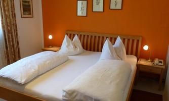 A cozy bedroom with a wooden bed and light cushions. The walls are painted orange, and there is soft lighting from bedside lamps.