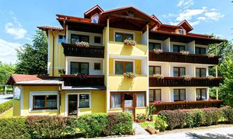 A bright yellow building with several floors and balconies, adorned with blooming plants. Surrounded by trees and a well-kept garden.