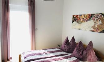 A bright room with a double bed and striped bed covers. A piece of artwork hangs on the wall and the windows are decorated with curtains made of purple fabric.