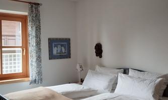 A cozy bedroom with a large double bed and a window. The walls are painted in light tones and adorned with some decorative elements.