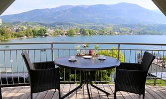 A beautiful view of a quiet lake with green hills and mountains in the background. Two chairs and a table are set up for a relaxed time outdoors.