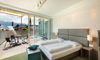 A modern bedroom with a double bed and a seating area. From the balcony, there is a view of the surroundings and the mountains.