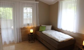 A bright room with a single bed, a small nightstand, and large windows. The soft curtains let in plenty of light and create a pleasant atmosphere.