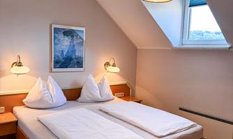 A cozy bedroom with a double bed and two pillows. In the room, there is a reading lamp, a picture on the wall, and a window with natural light.
