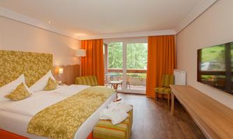 A modern hotel room with a large bed and orange curtains. There is a television and a balcony overlooking nature.