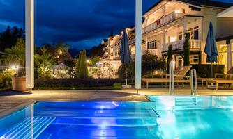 An elegant outdoor pool with blue light at night, surrounded by stylish architecture. In the background, gardens and terraces are visible.