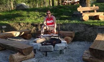A cozy fire pit in nature with wooden benches and a grill. In the background, there are trees and a rustic setting.