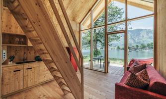 A modern wooden house with a large window front that opens up the view of a lake. Inside, a staircase and a cozy seating area can be seen.