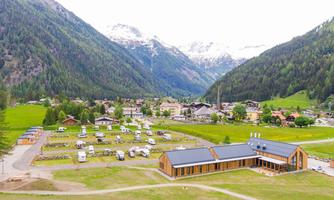 A picturesque valley with green meadows and mountains in the background. In the foreground are motorhomes and a modern building.