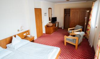 A simple hotel room with a double bed, two chairs, and a table. The room has carpeted flooring and large windows for natural light.