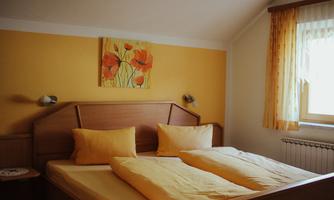 A cozy bedroom with a large double bed and yellow cushions. The walls are painted in a warm shade of yellow, and a painting with flowers adorns the wall.