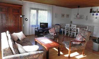 A cozy living room with a sofa, two armchairs, and a table. In the background, there is a dining table and a television in front of a window.