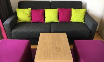 A gray sofa with colorful cushions in pink and green. Alongside it is a square wooden table and two square, pink stools.