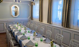 An elegant dining room with a long table, set with white tablecloths and blue accents. The walls are painted in light colors and large windows provide plenty of light.