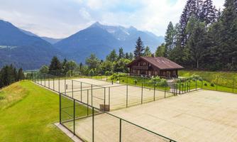 A picturesque tennis facility in the mountains with several tennis courts and a cozy wooden house in the background. The surroundings are characterized by green meadows and tall trees.