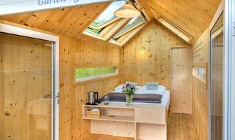 A cozy room made of wood with a window and a glass door. In the room, there is a bed, a table with a coffee pot, and fresh flowers.