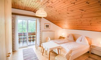 A cozy bedroom with a wooden ceiling and two beds. Large windows offer a view of the green surroundings.