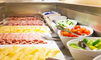 A selection of sausages and cheese is placed on a tray, complemented by fresh vegetables and tomatoes in bowls. The image showcases an appetizing breakfast bar.