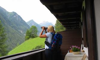 A woman stands on a balcony and looks at the mountains with binoculars. In the background, green hills and wooded landscapes are visible.