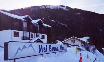 A winter view with snow-covered hills and buildings. The sign "Mai Brunn Alm" is clearly visible.