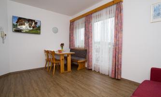 A cozy living space with a dining table, a comfortable seating area, and a television on the wall. Large windows with curtains provide plenty of light.