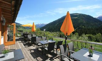 A sunny terrace with tables and chairs, surrounded by green mountains. Orange umbrellas provide shade for the guests.