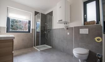 A modern bathroom with a shower and a toilet. It has large windows and a bright, friendly impression.