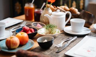 A set dining table with a selection of breakfast ingredients, including fresh fruit, cold cuts, and beverages. A light teapot and a vase with orange flowers complete the picture.