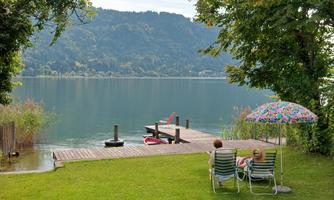 A calm lake with a wooden pier and green meadow. Two people are relaxing under a colorful sunshade.