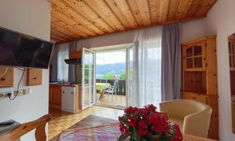 A cozy living and dining room with a wooden ceiling and a large window that opens onto a balcony with a view. A blooming floral arrangement is on the table.
