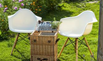 Two elegant white chairs stand on a green lawn, surrounded by blooming flowers. Next to the chairs is a woven basket with plants and garden tools.