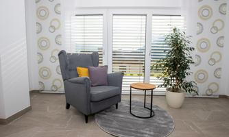 A cozy armchair with cushions stands next to a small table in a bright room. Large windows let in plenty of light, and a plant creates a pleasant atmosphere.
